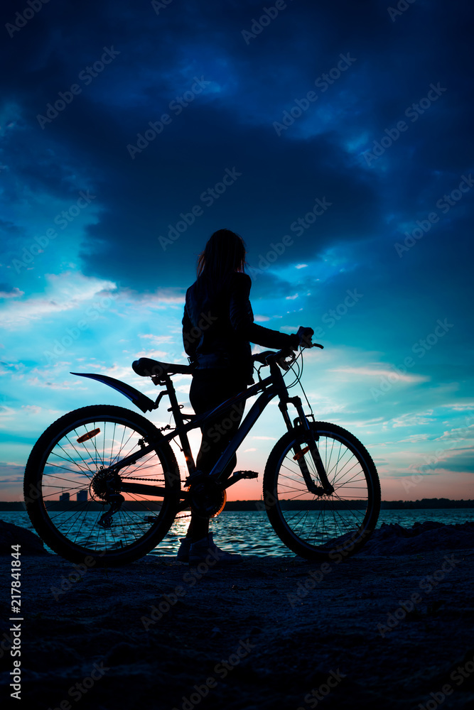 Fototapeta premium Silhouette of a young woman with a bicycle on the lake at sunset