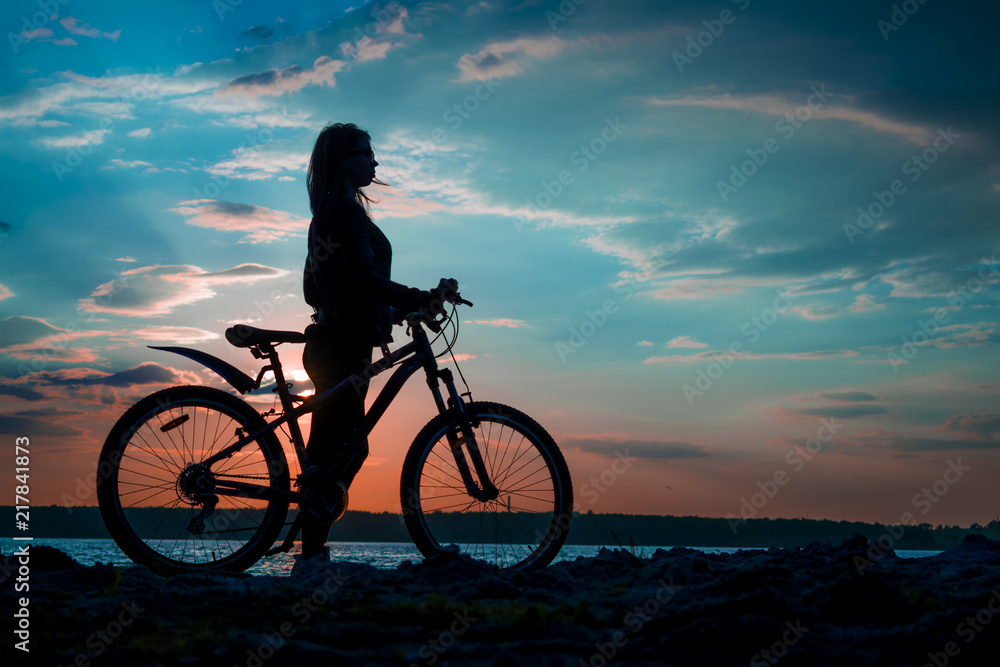 Fototapeta premium Silhouette of a young woman with a bicycle on the lake at sunset