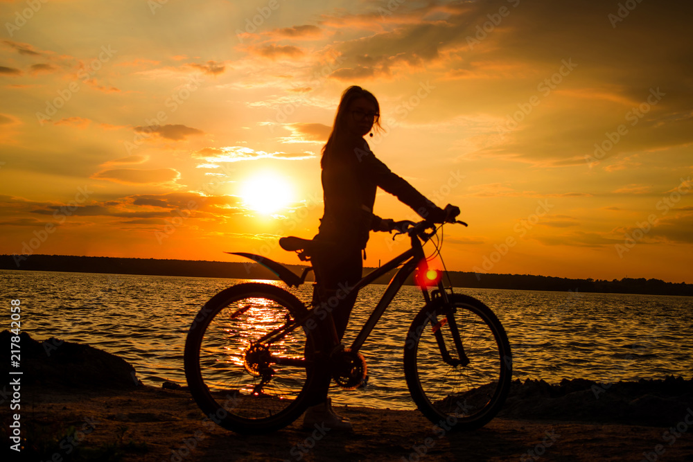 Fototapeta premium Silhouette of a young woman with a bicycle on the lake at sunset