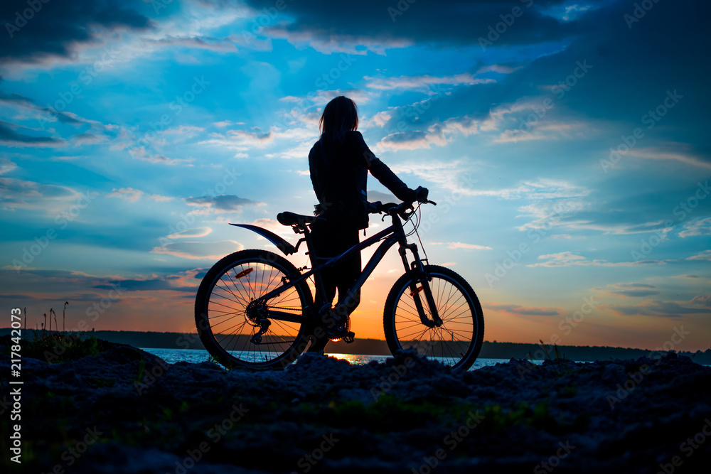 Fototapeta premium Silhouette of a young woman with a bicycle on the lake at sunset