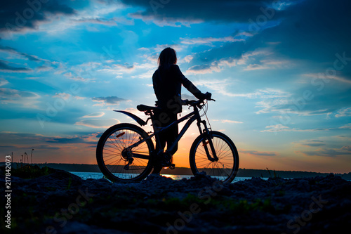 Silhouette of a young woman with a bicycle on the lake at sunset