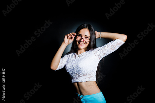 Portrait of an attractive young woman in white top and blue pants posing with her glasses in the dark.