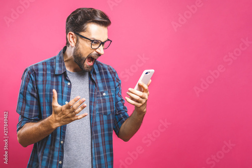 Young caucasian man angry, frustrated and furious with his phone, angry with customer service over pink background. photo