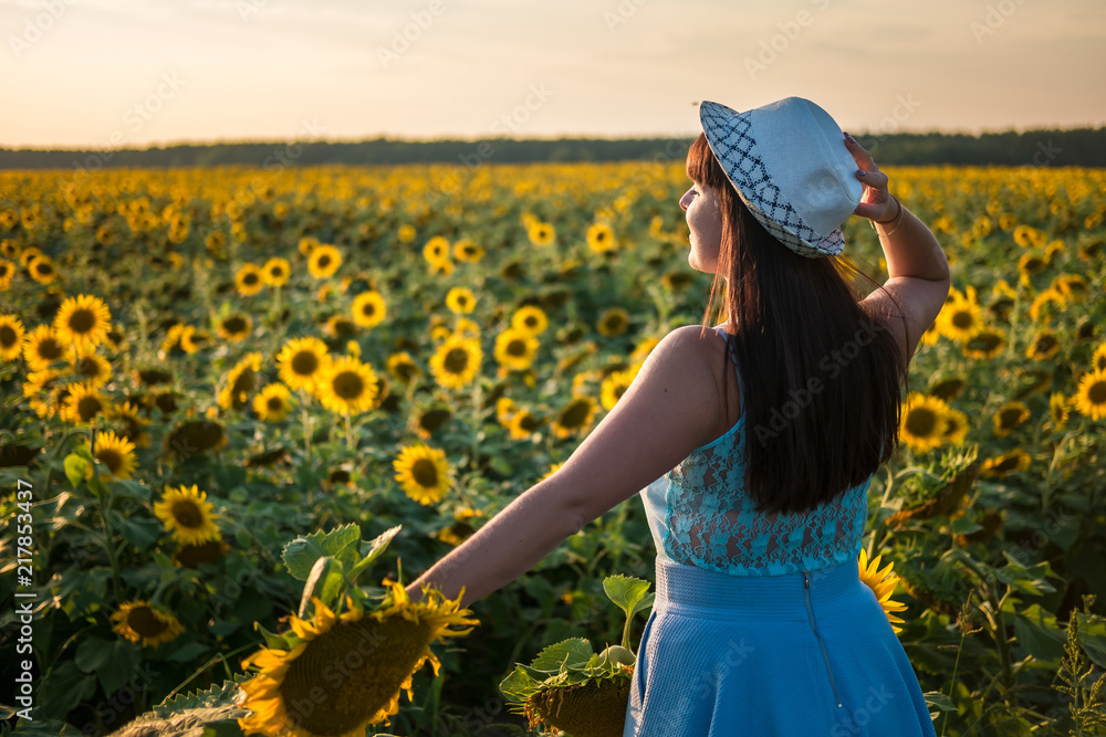 Girl in a blue dress leaves with hat in field of sunflowers on the sunset. Follow me concept