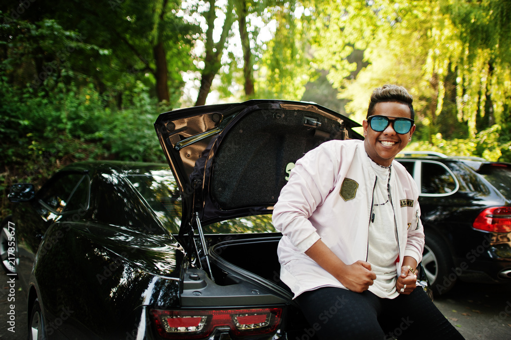 Stylish hipster arab man guy in sunglasses posed outdoor in street and sitting on the trunk his black muscle car. Rich black man.