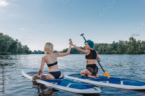 Canvas Print athletic women giving highfive while sitting on paddle boards on river