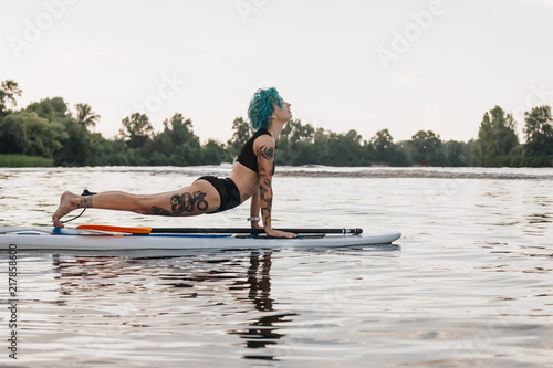 tattooed woman with blue hair practicing yoga on paddleboard in water. Upward facing dog pose  Urdhva Mukha Svanasana 