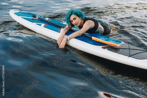 beautiful athletic tattooed young woman with blue hair relaxing on sup board in water