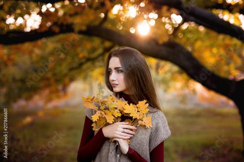 Seasonal autumn fashion portrait. Modern young woman wearing fashionable warm clothes posing in the autumn park holding yellow leaves.