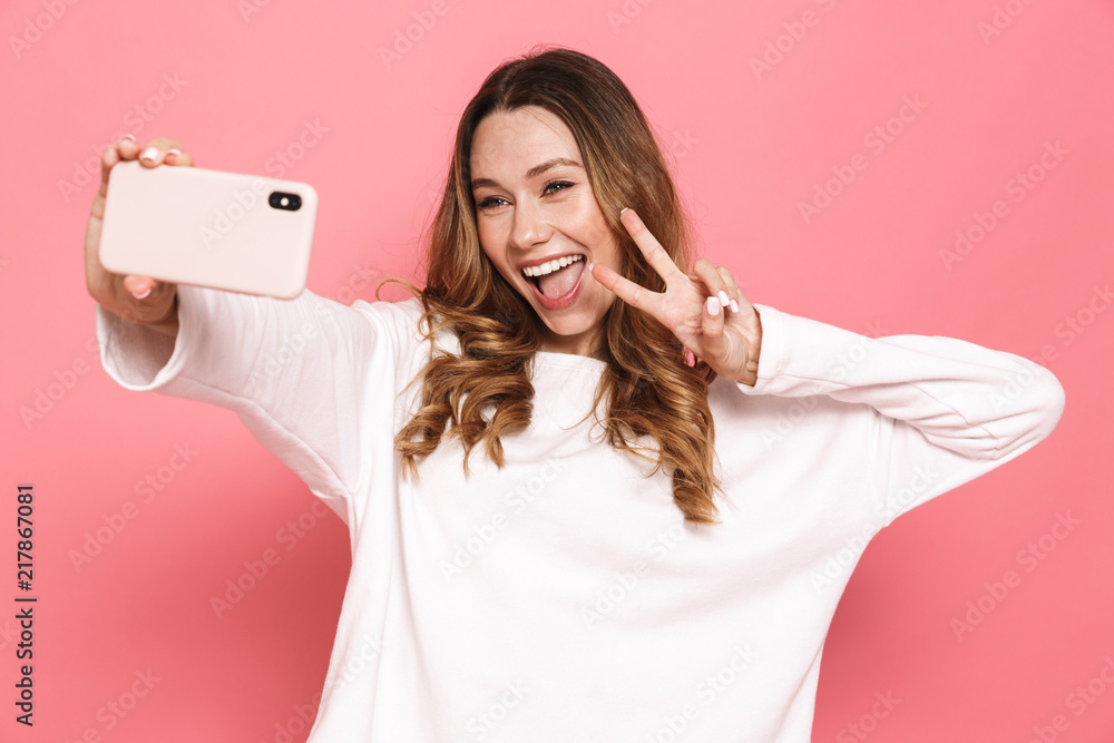 Obraz premium Photo of cheerful brunette woman with beautiful long hair showing peace sign and taking selfie on cell phone, isolated over pink background