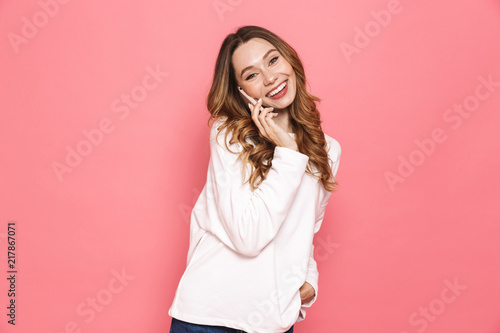 Image of smiling brunette woman with beautiful long hair holding cell phone in hands and having mobile conversation, isolated over pink background