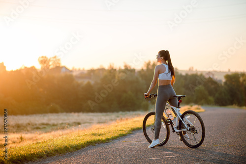 Rear view of young brunette fit woman in sportswear with bicycle on the countryside road near the field on sunset.