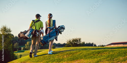 Full length of a happy couple with a healthy lifestyle wearing golf outfits, while carrying stand bags with professional clubs towards the golf course in a sunny day of summer photo