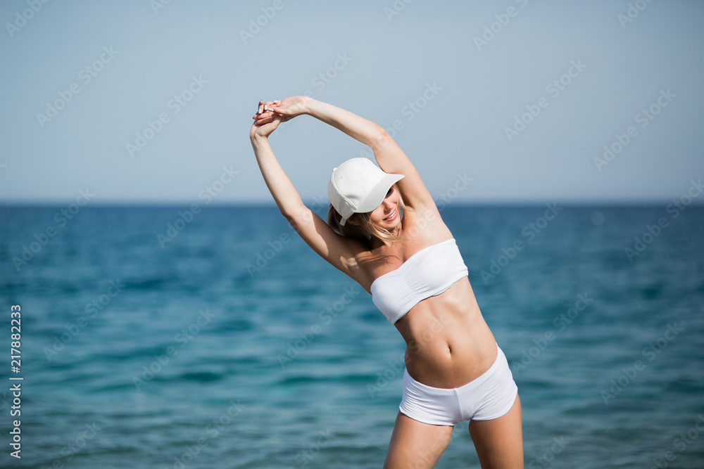 Fototapeta premium Slim female girl with beautiful figure is doing stretching exercise on the beach, morning run, fitness and healthy lifestyle concept