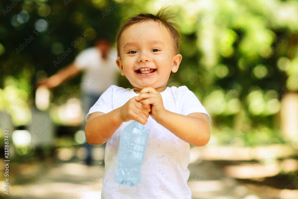 Laughing little boy running outdoor with a bottle of a clear drinking water, his father is blurred in the background. Importance of clear water concept Laughing little boy running outdoor with a bottle of a clear drinking water, his father is blurred in the background. Importance of clear water concept