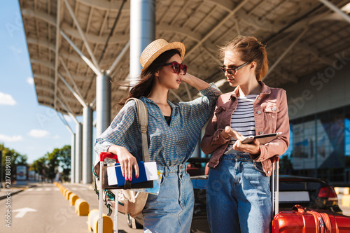 Young female friends in sunglasses amazedly looking at each other while holding passport with tickets and tablet in hands with airport on background
