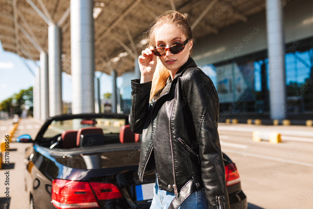 Beautiful girl in sunglasses and leather jacket thoughtfully looking in camera near airport with black cabriolet on background