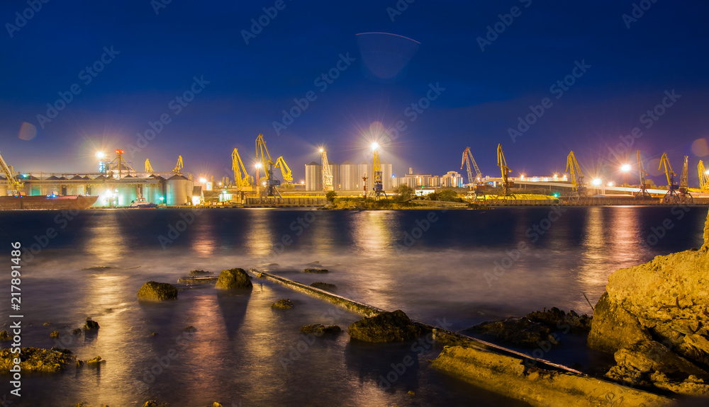 Obraz premium Night view of an estuary with a port and cranes and ships on the other shore. Seascape.