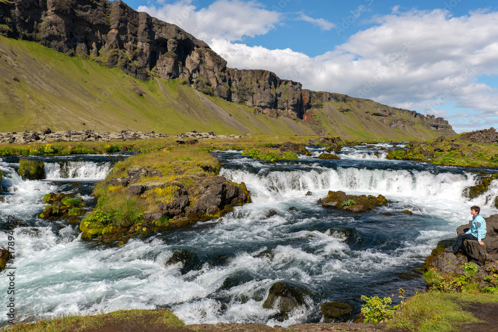 Urlaub in Island, Frau sitzt an Flusslandschaft mit kleinen Wassertreppen