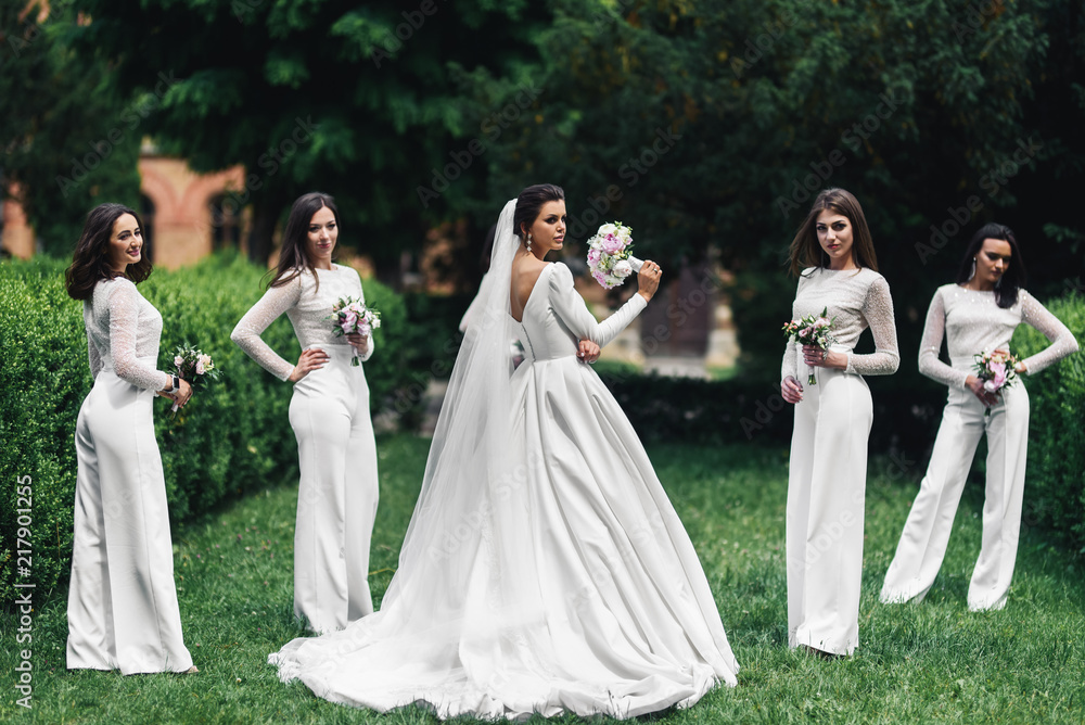 Beautiful bride and bridesmaids with wedding bouquets pose on the green lawn