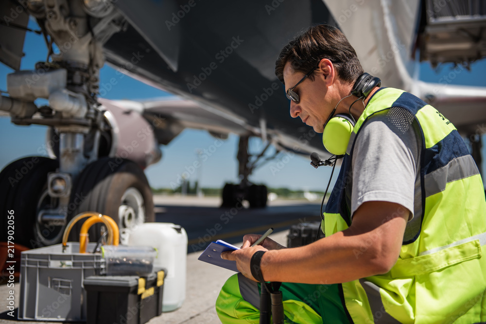 Fototapeta premium Important information. Ground crew member sitting on haunches while noting data. Airplane landing gear on background
