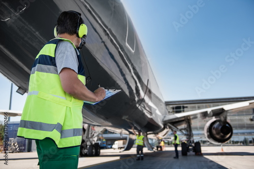 Good job. Back view of man in headphones noting data while observing aircraft. Ground crew members preparing plane for the flight in the background