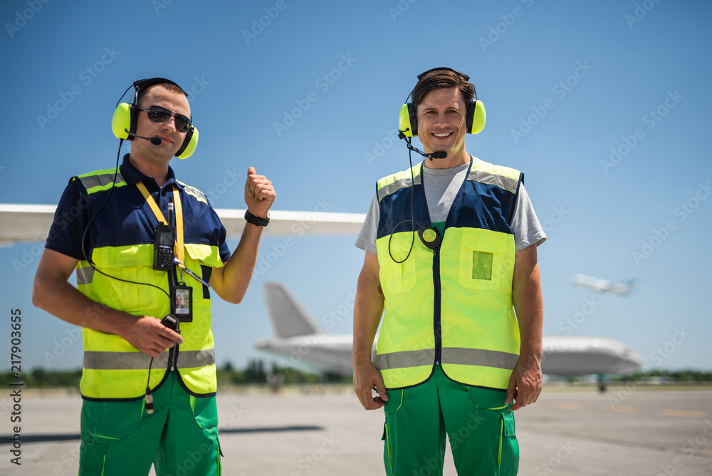 Feeling joyful. Smiling man posing at airdrome while colleague looking at camera with raised hand. Blue sky, runway and planes on blurred background Feeling joyful. Smiling man posing at airdrome while colleague looking at camera with raised hand. Blue sky, runway and planes on blurred background