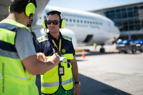 Right here. Smiling man in headphones pointing at clipboard while partner looking at writing data. Passenger plane on blurred background