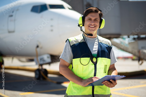 Time for fun. Waist up portrait of smiling worker at airport. Man holding clipboard and wearing headphones with microphone