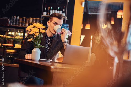 Handsome freelancer man with stylish beard and hair dressed in a black suit sitting at a cafe with an open laptop and drinks a coffee  looking at the camera.