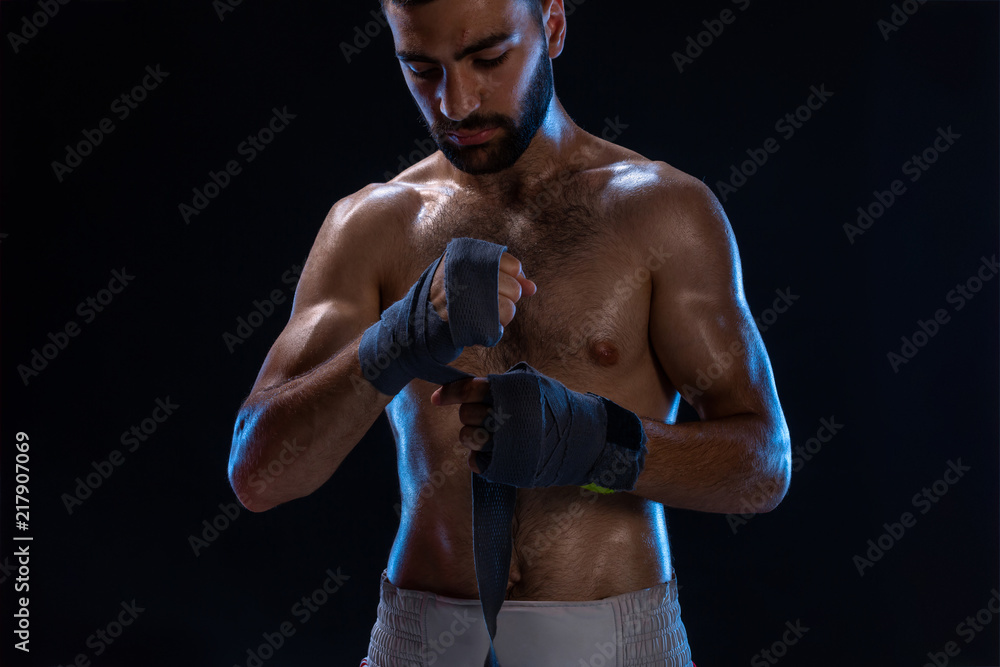 Sports boxer man pulls on the hand wrist wraps. Oriental male model isolated on black background.