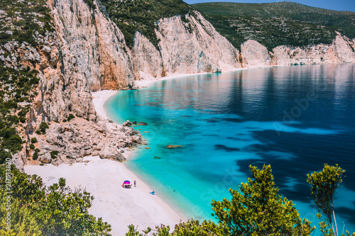Amazing Fteri beach lagoon, Kefalonia, Greece. Tourists under umbrella chill relax near clear blue emerald turquoise sea water. Cliff rock coastline in background