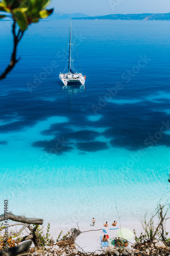 Fototapeta Naklejka Na Ścianę i Meble -  White catamaran yacht at anchor on clear azure surface with dark pattern in calm blue lagoon. Unrecognizable tourists relax under sun umbrella on the beach