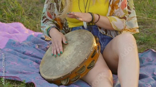 young blond hippie woman sitting on grass in park at sunset. Warm summer day and soft backlight. happy woman playing the tambourine and singing. slow motion HD photo