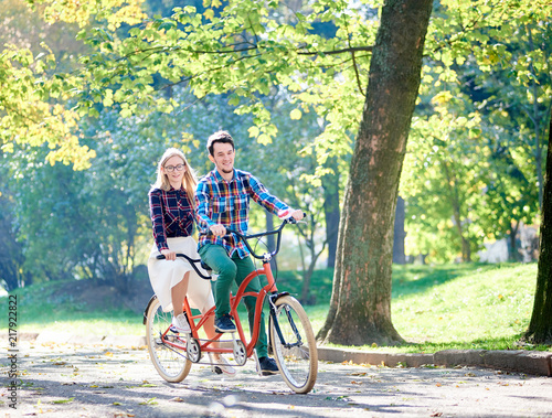 Young active smiling traveler couple, handsome bearded man and attractive blond woman cycling together tandem double bike along crackled path in lit by bright sun beautiful park under tall trees.