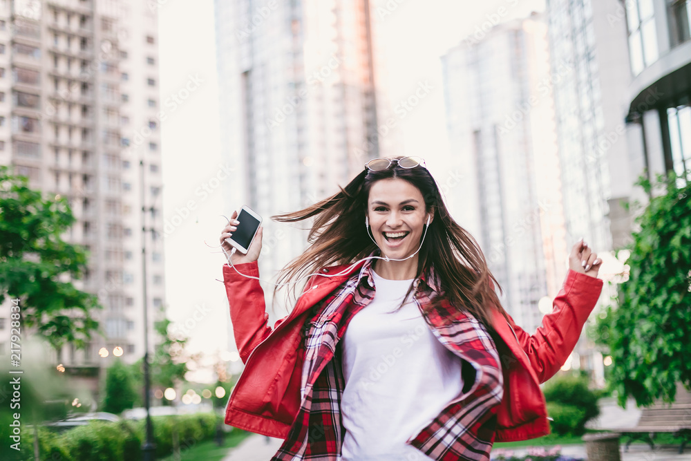 Beautiful young expressive woman in red jacket with mobile phone on modern city street, she is happy, enjoying of music in headphones.