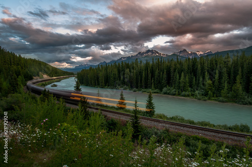 Train rushing by river and forest with Rocky Mountains in background at sunset