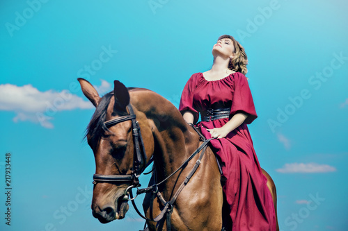 A young woman sits in a red dress is sitting on the back of a brown horse against a blue sky and clouds.