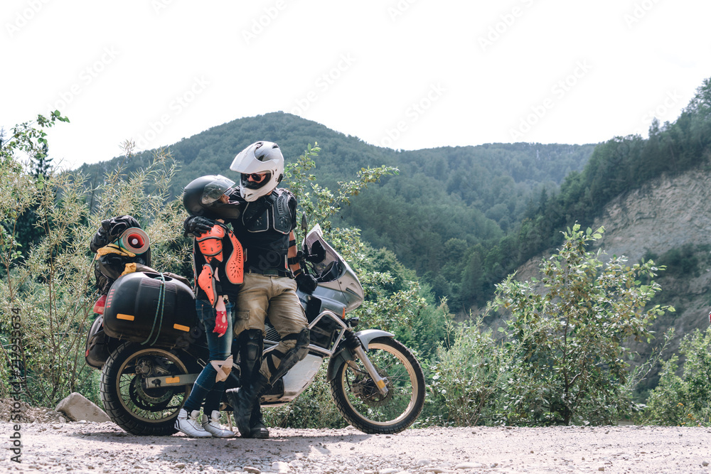 Happy young love couple in body armor and halmets stand with big adventure motorcycle, travel togather, jorney, copy spase, mountains dirt road, enduro road trip Happy young love couple in body armor and halmets stand with big adventure motorcycle, travel togather, jorney, copy spase, mountains dirt road, enduro road trip