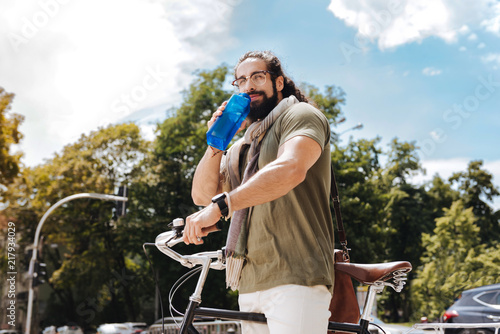 Fresh water. Nice delighted man standing near his bicycle while drinking water