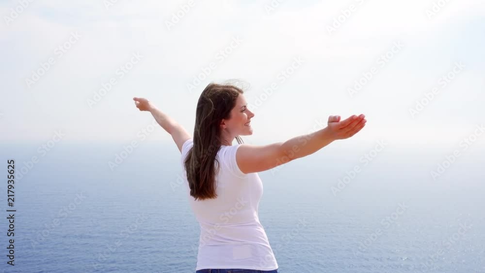 Smiling young woman in white t-shirt raising arms up at cliff against breathtaking view of blue sea. Carefree female outstretching hands. Concept of freedom and inspiration Smiling young woman in white t-shirt raising arms up at cliff against breathtaking view of blue sea. Carefree female outstretching hands. Concept of freedom and inspiration
