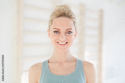 Young slim woman in gray top standing in light white hall and smiling tenderly at camera