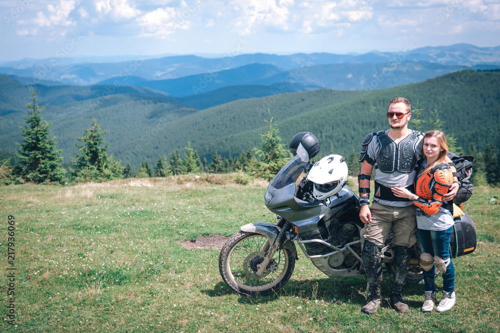 Obraz premium Happy young love couple in body armor and halmets stand on the top of the mountain, with big adventure motorcycle, travel togather, jorney, copy spase, mountains dirt road, enduro road trip