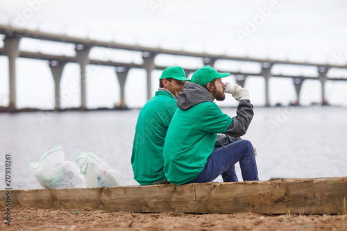 Side view of volunteers in green uniform sitting with coffee cups and relaxing on shore