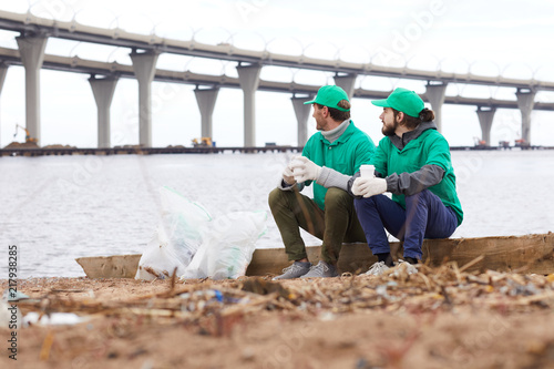 Volunteers in green uniform sitting with paper cups and looking away while resting on shore