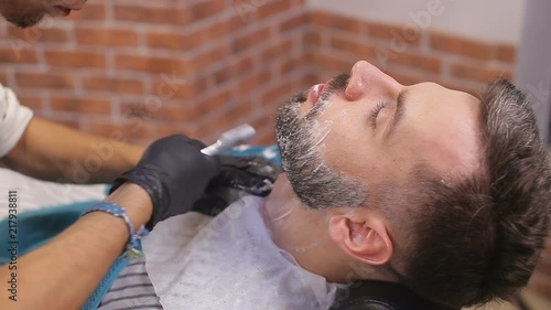 Bearded male sitting in an armchair in a barber shop while hairdresser shaves his beard with a dangerous razor photo