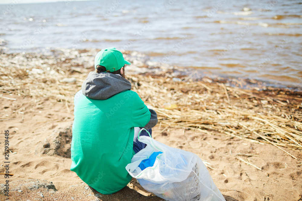 Back view of volunteer in green uniform sitting on shore at plastic bag and looking away Back view of volunteer in green uniform sitting on shore at plastic bag and looking away