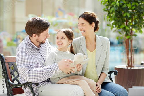 Happy parents with charming girl sitting on bench in light shopping center having fun and playing with toy