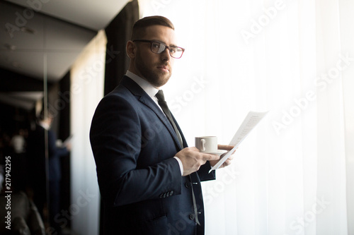 Adult serious man in elegant suit and eyeglasses holding cup of coffee and paper documents in room looking at camera