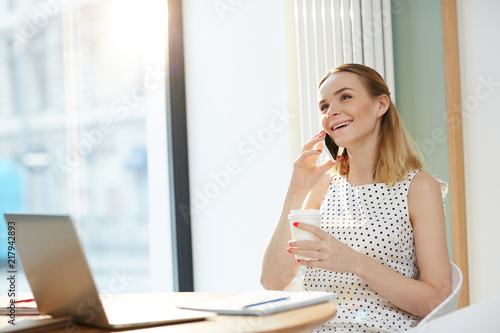 Relaxed modern businesswoman holding paper cup of coffee and smiling while speaking on smartphone at table with laptop